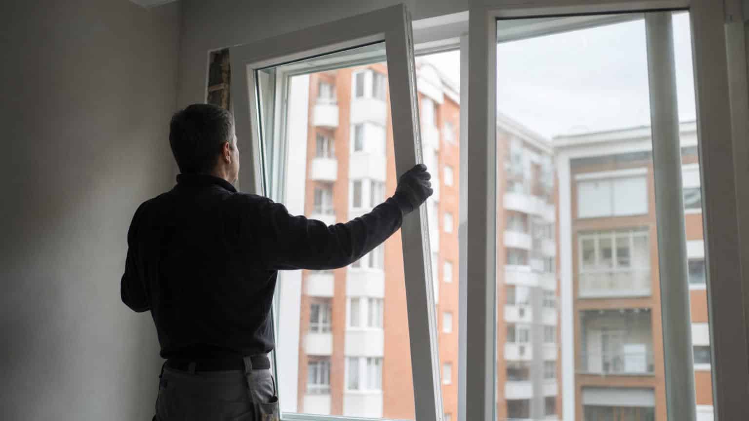 Worker installing a new white PVC window frame in an apartment overlooking urban brick buildings.